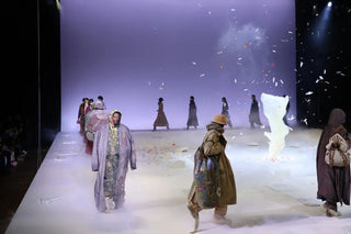 Models walking on foggy white runway with oversized coats and hats during fashion show with floral confetti