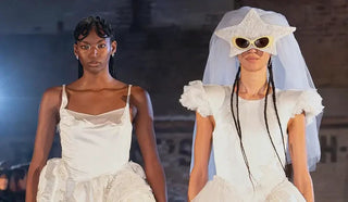 Two models wearing white avant-garde bridal-inspired dresses on runway, one with star-shaped sunglasses and veil