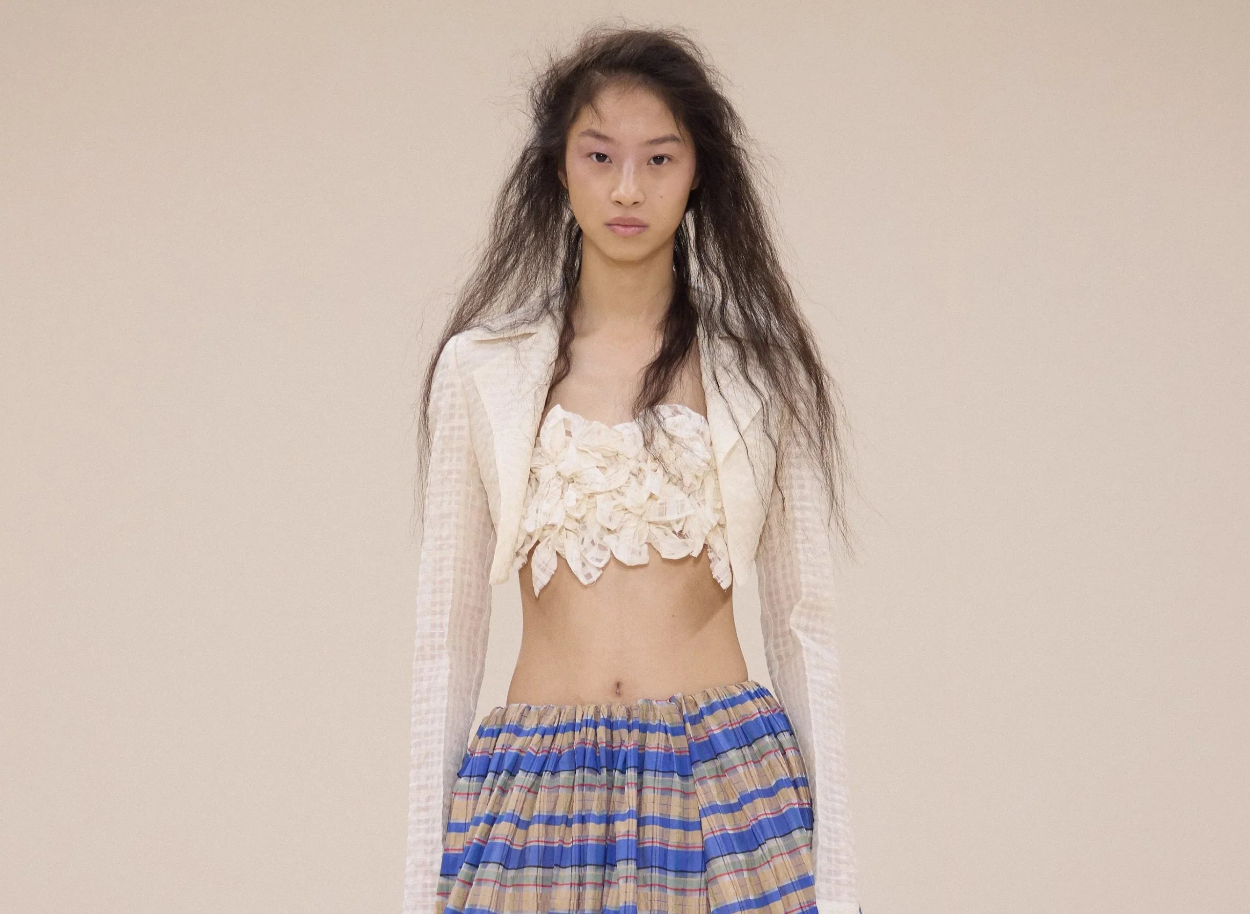 Fashion model with long textured hair wearing a sheer white cropped jacket, lace top, and striped skirt