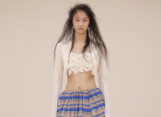 Fashion model with long textured hair wearing a sheer white cropped jacket, lace top, and striped skirt