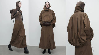 Model wearing brown hooded jacket and long skirt with black boots, carrying dark brown bag in minimalist setting