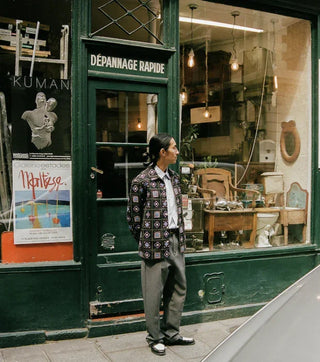 Man in patterned jacket and gray pants standing outside a vintage green storefront with Dépannage Rapide sign