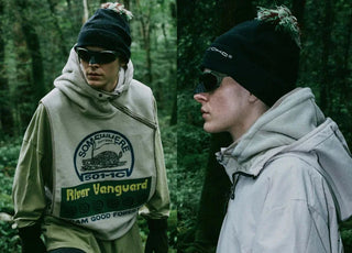 Two men wearing outdoor hoodies, black beanies, and sunglasses in a forest setting