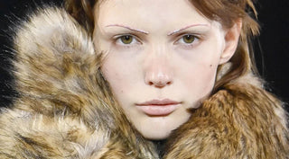 Close-up of a female model with minimal makeup, thin styled eyebrows, and a large brown faux fur collar