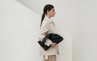 Woman in beige trench coat holding large black clutch bag against white background