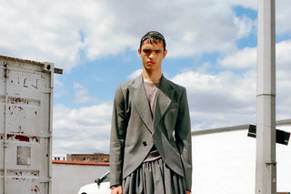 Young man in gray blazer and skirt standing outdoors under cloudy sky with urban background