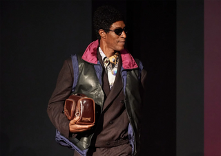 Fashion model wearing layered outfit and sunglasses holding a brown leather Martine bag on runway