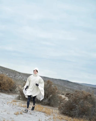 Person running in a white rain poncho and black gloves in a dry desert landscape with cloudy sky