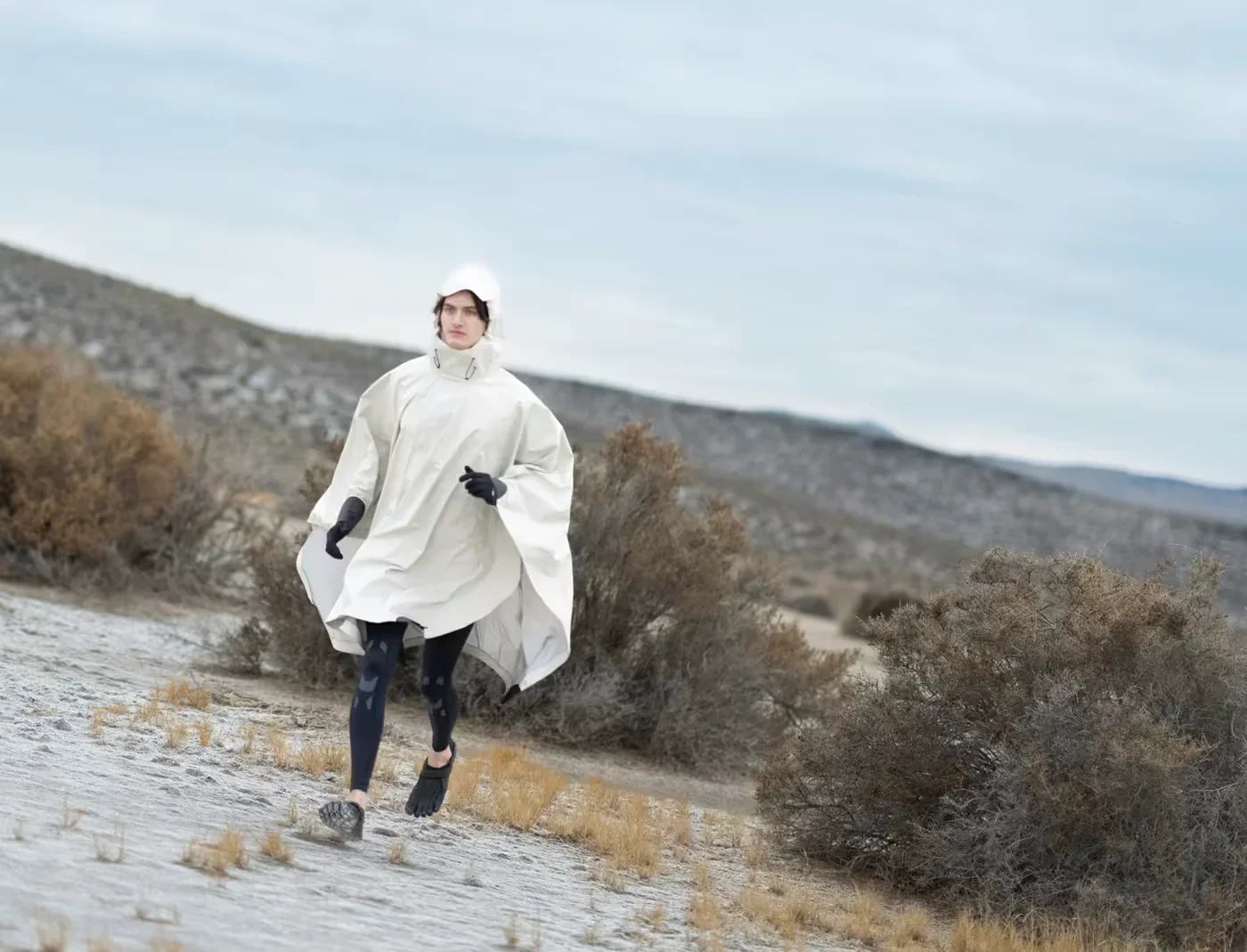 Person wearing white hooded rain poncho and black leggings walking in dry desert landscape with shrubs