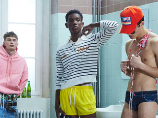 Three male models in a bathroom wearing colorful streetwear and swimwear, featuring hoodies, striped tops, and swim trunks