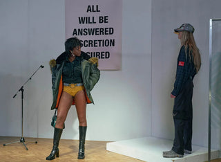 Two models at fashion event, one in green parka and yellow shorts, other in plaid shirt and black pants