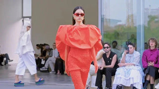 Model wearing bold red avant-garde outfit and sunglasses walking on runway with seated audience in modern fashion show