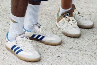 Two pairs of feet wearing vintage white Adidas sneakers with blue and brown accents on concrete
