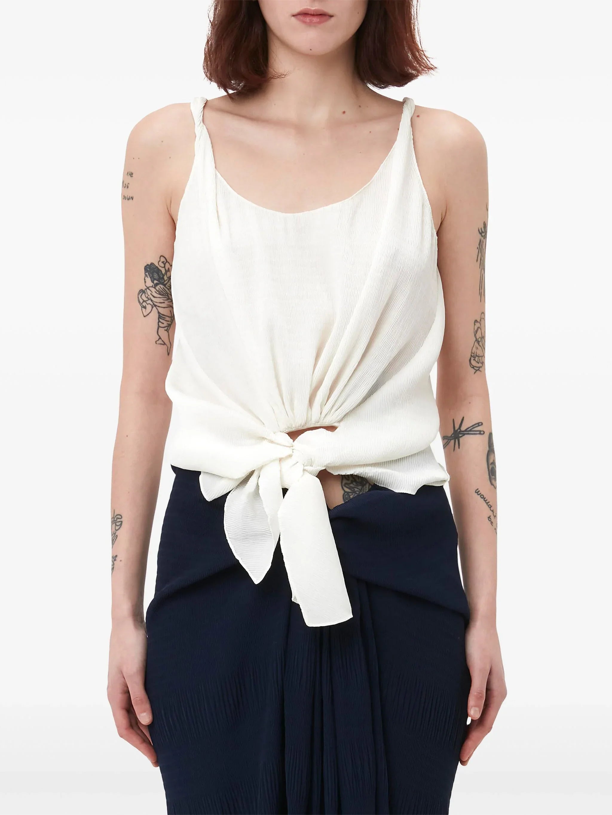 Woman wearing white sleeveless tie-front top and dark pleated skirt with tattoos on arms