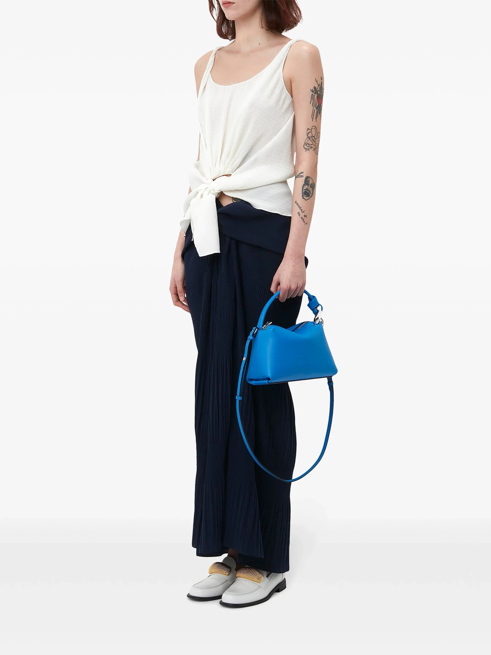 Woman in white sleeveless top and navy skirt holding blue leather handbag with tattoos on arm