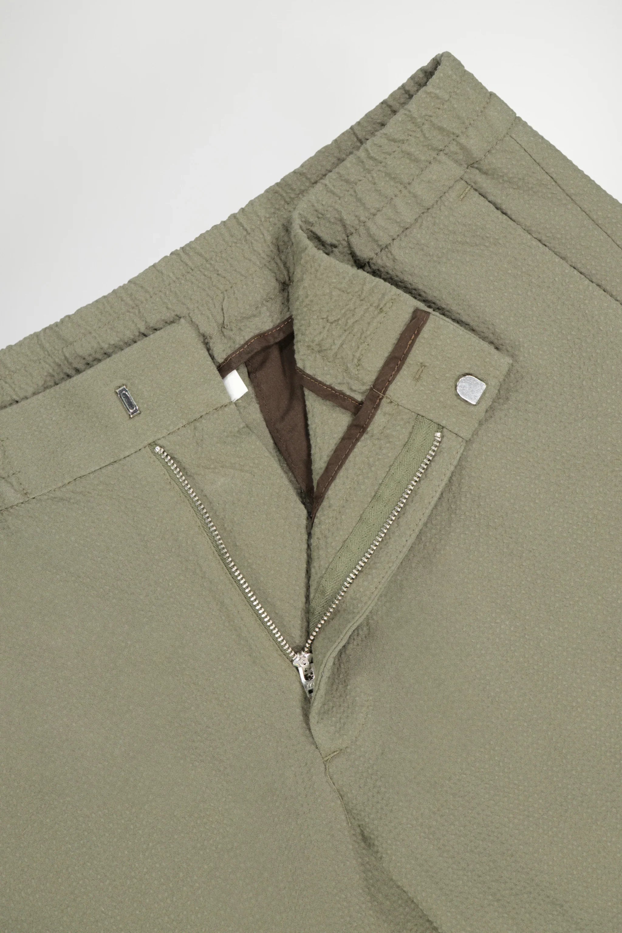 Close-up of olive green textured pants with elastic waistband, zipper fly, and snap button closure