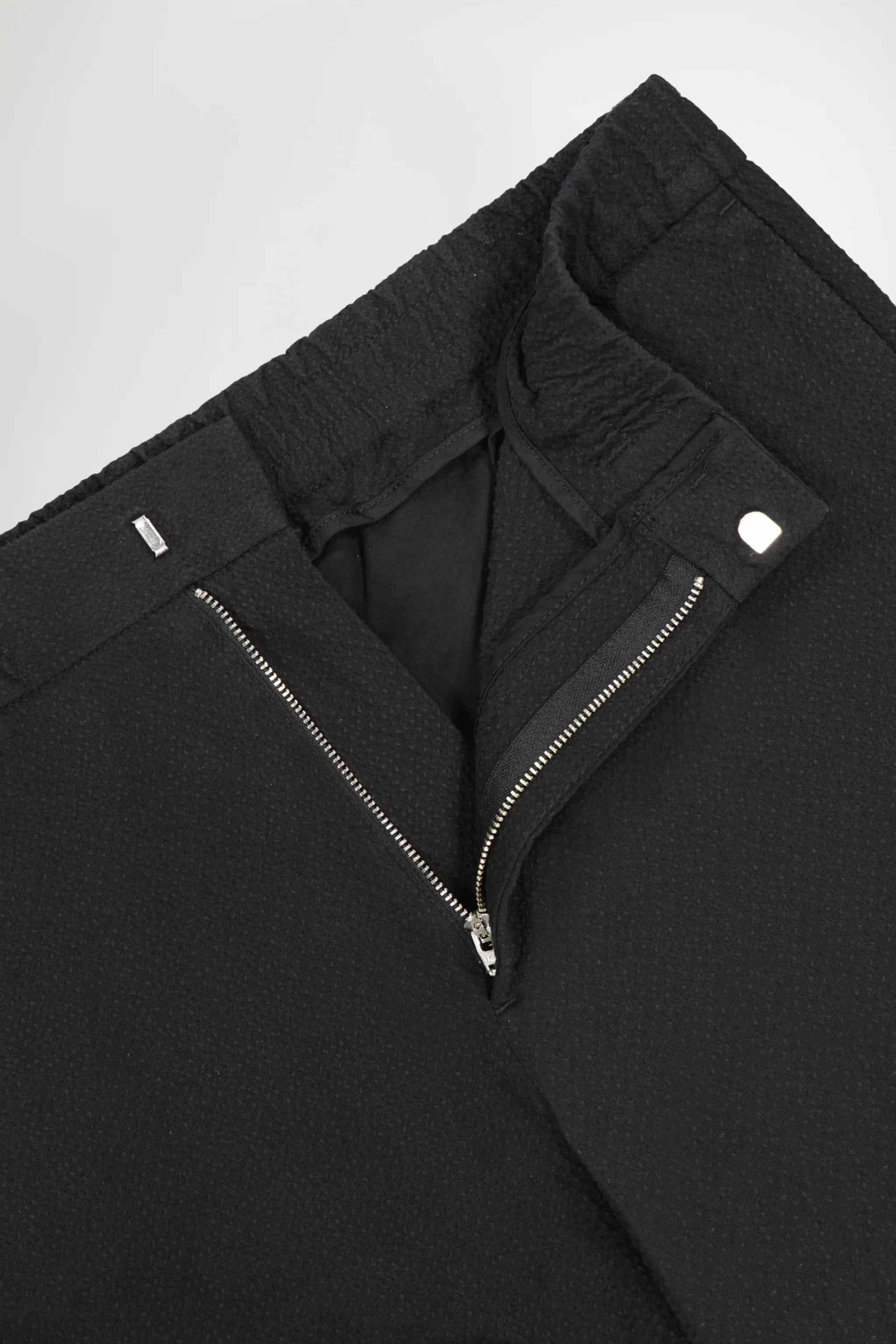 Close-up of black textured pants with silver zipper and button closure on white background