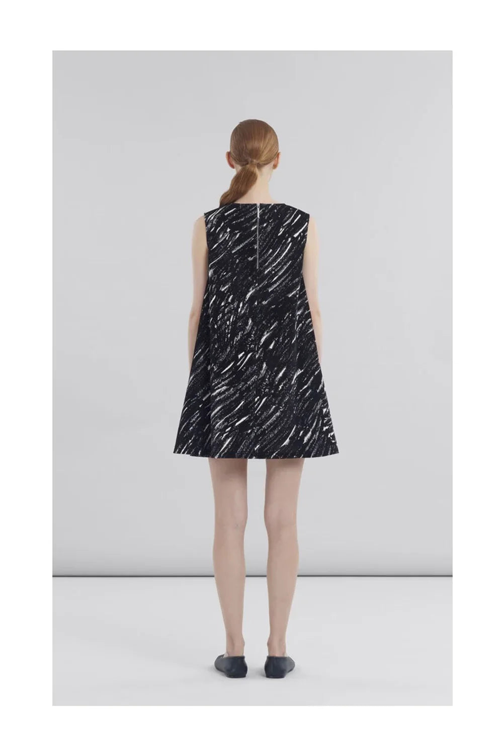 Back view of woman wearing sleeveless black and white abstract print A-line dress with black shoes on plain background