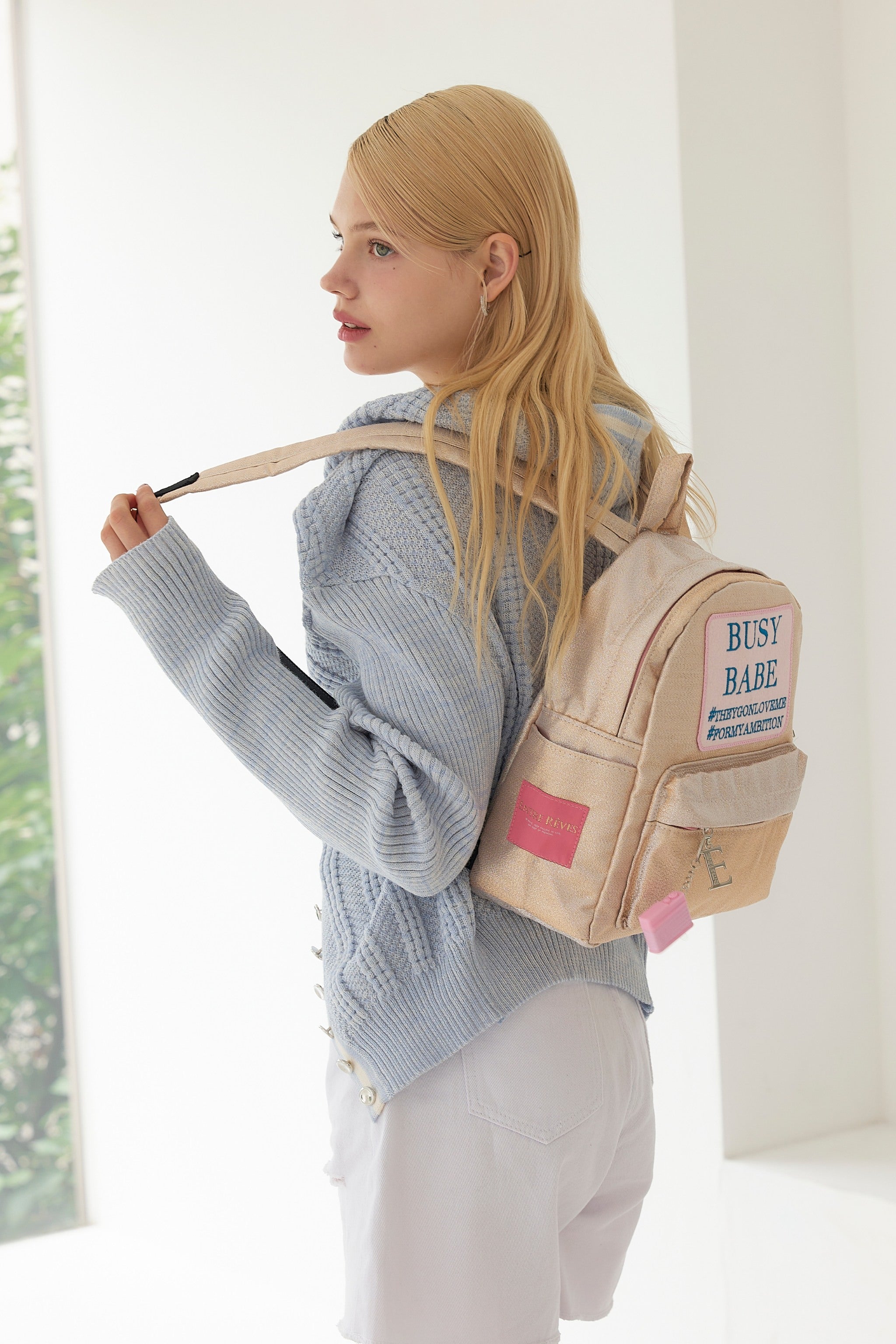 Woman wearing a beige backpack with text, standing indoors.
ENTRE REVES - Busy Babe Gold Mini beige backpack with a pink patch displaying text on a white background.