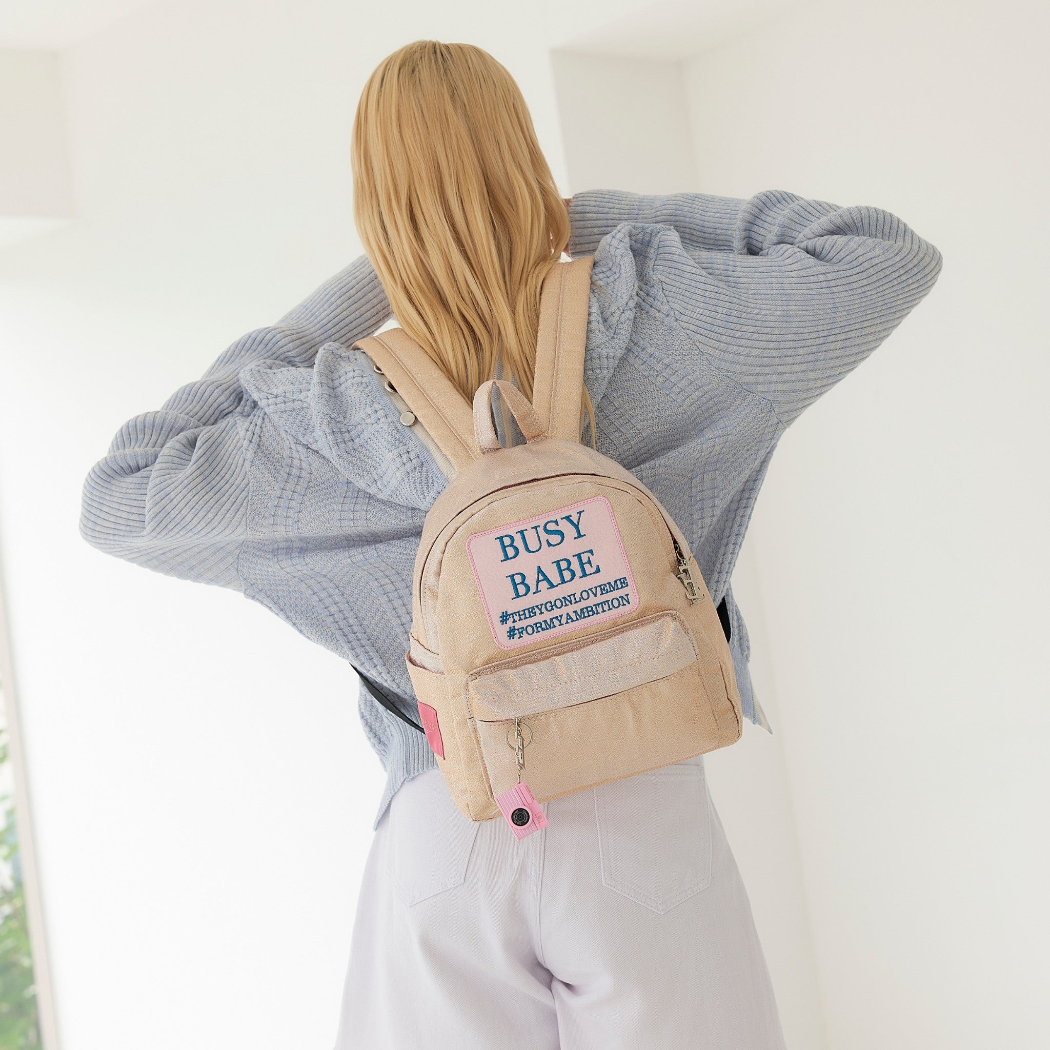 Woman wearing a beige backpack with text, standing indoors.
ENTRE REVES - Busy Babe Gold Mini beige backpack with a pink patch displaying text on a white background.