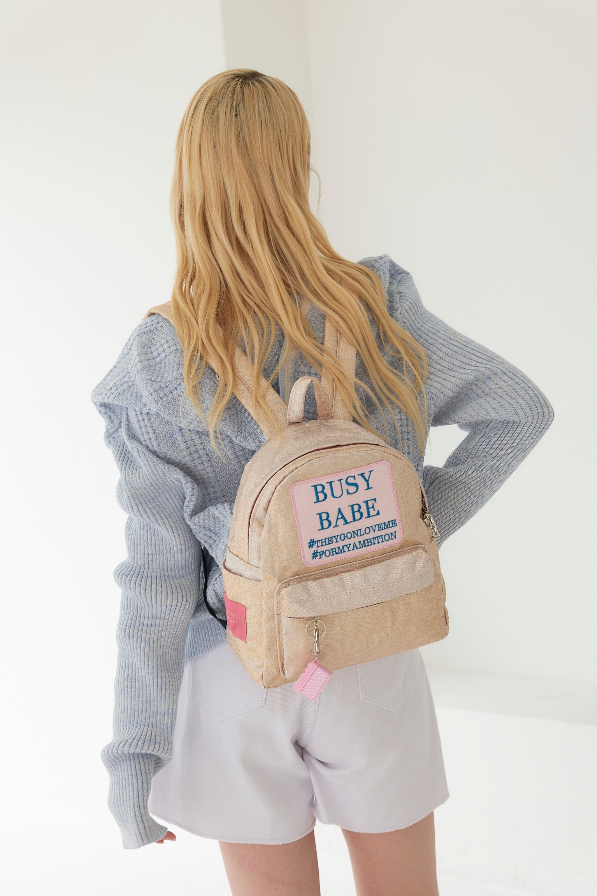Woman wearing a beige backpack with text, standing indoors.
ENTRE REVES - Busy Babe Gold Mini beige backpack with a pink patch displaying text on a white background.
