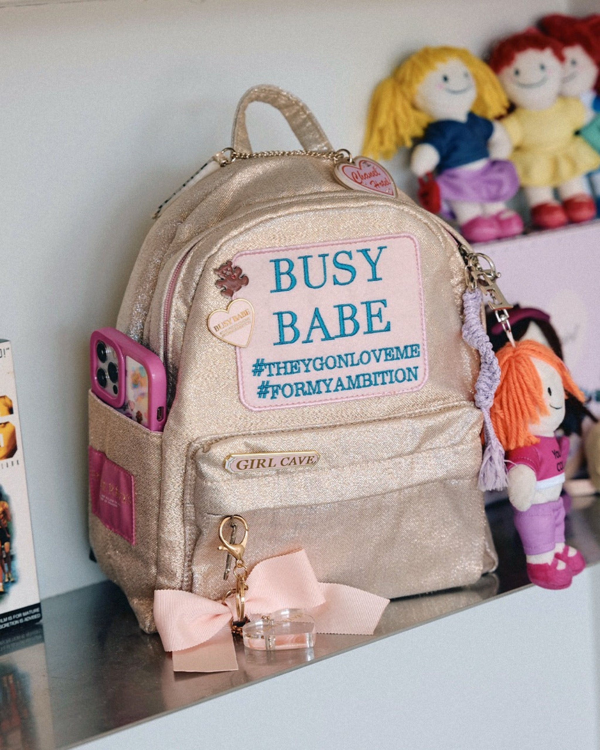 ENTRE REVES - Busy Babe Gold Mini beige backpack with a pink patch displaying text on a white background.
Gold backpack with 'Busy Babe' text on a shelf with toys in the background