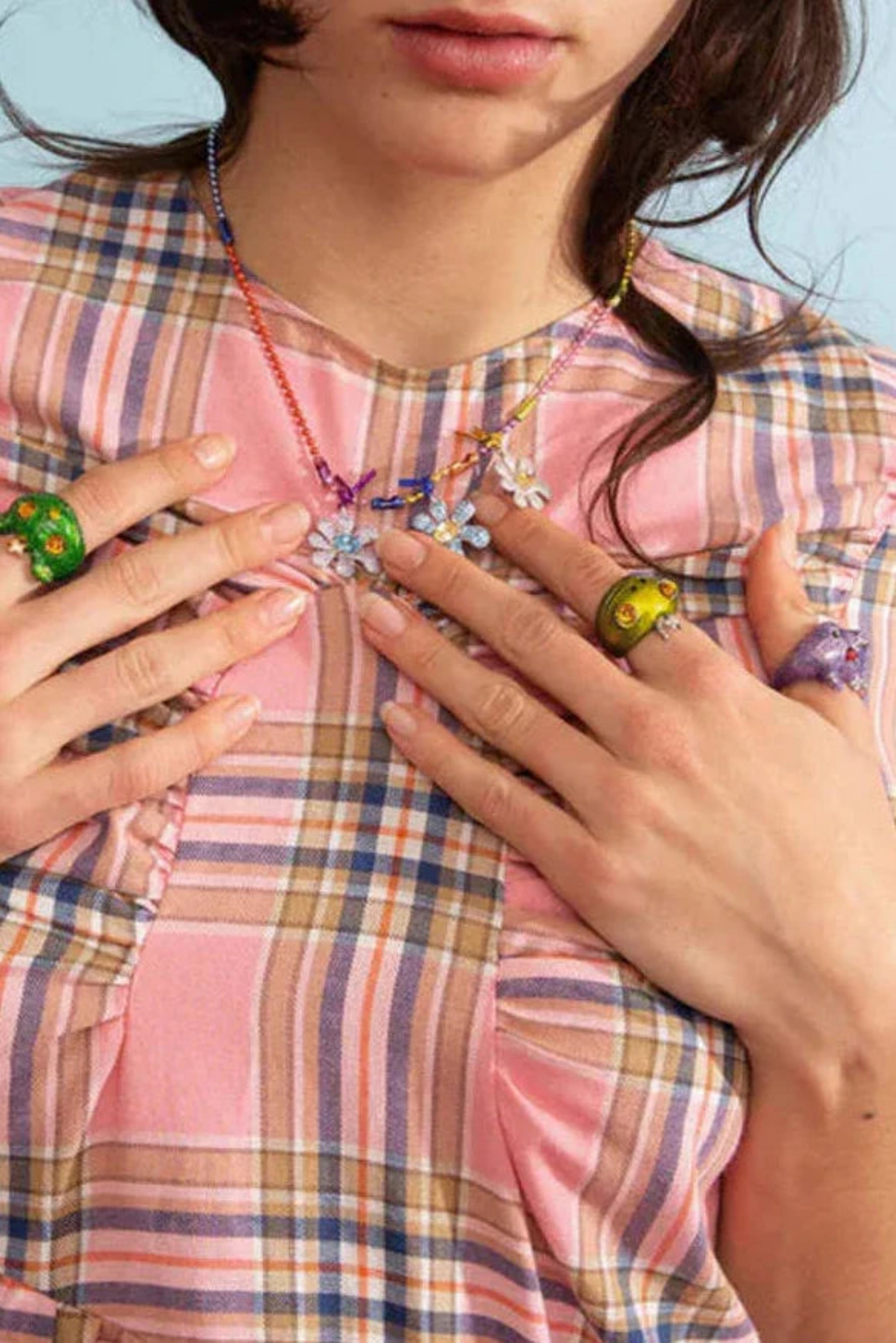 Close-up of a woman wearing colorful flower necklace and oversized green, yellow, purple rings on a pink plaid shirt