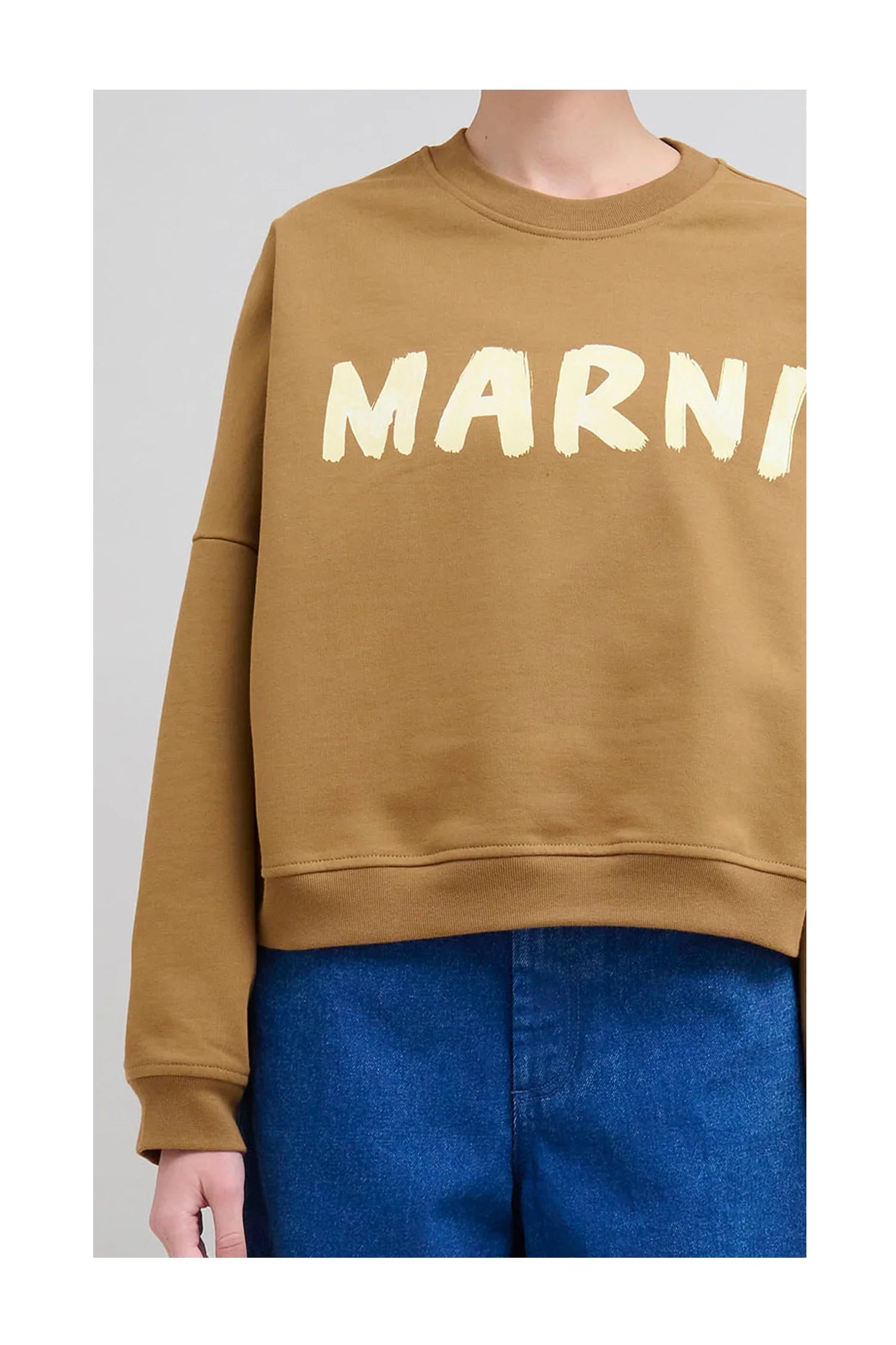 Close-up of person wearing brown sweatshirt with large light yellow Marni logo and blue denim jeans