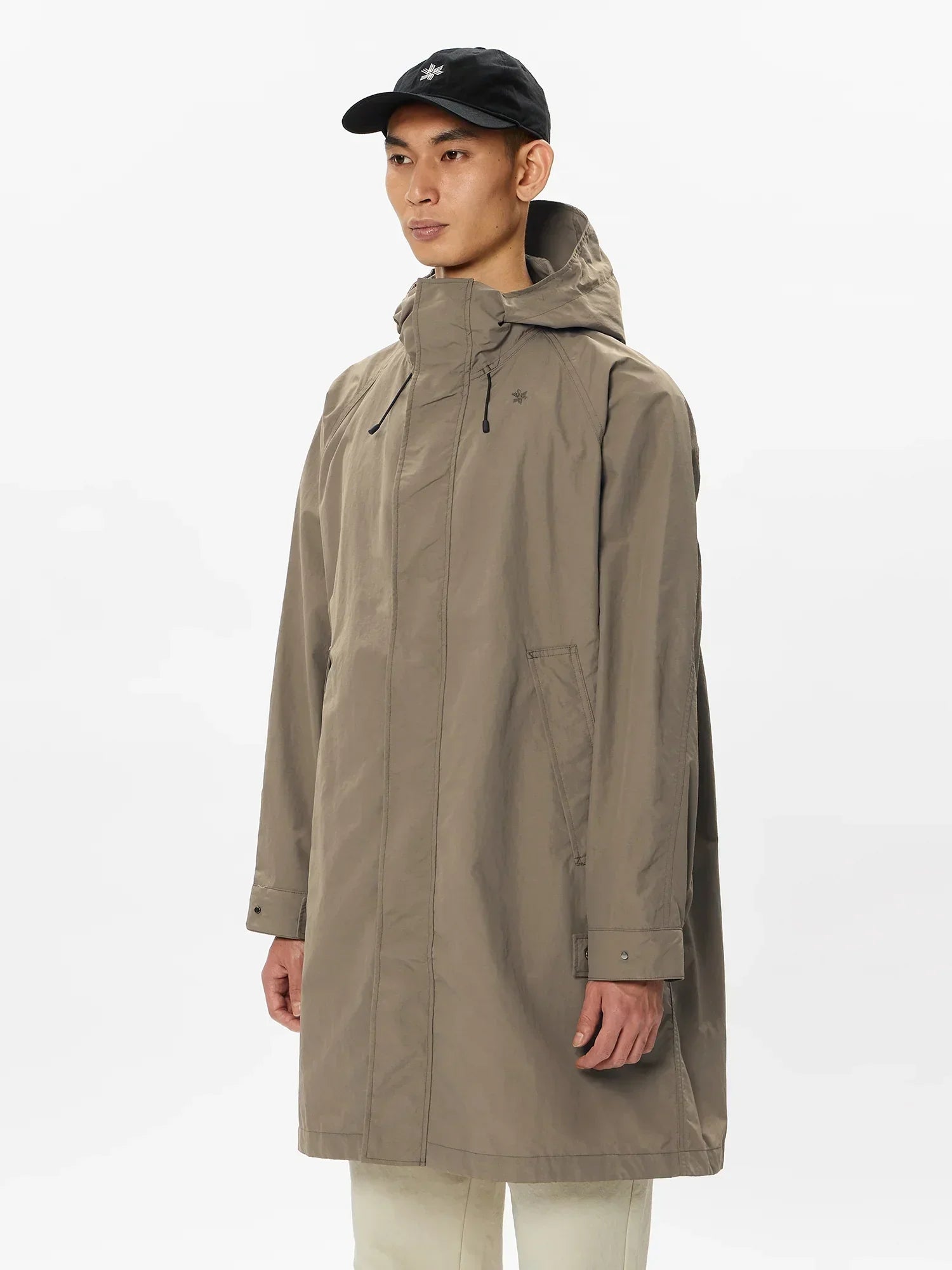 Man wearing beige hooded rain jacket with front pockets and black cap on white background