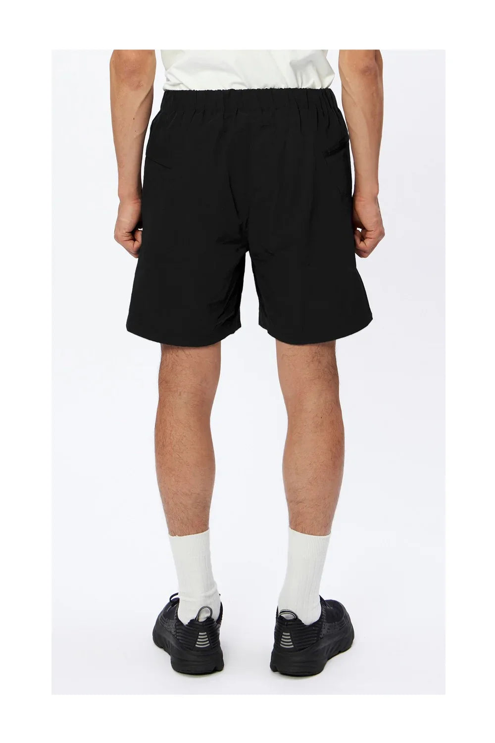Rear view of a man wearing black elastic waist shorts, white socks, and black sneakers on a white background