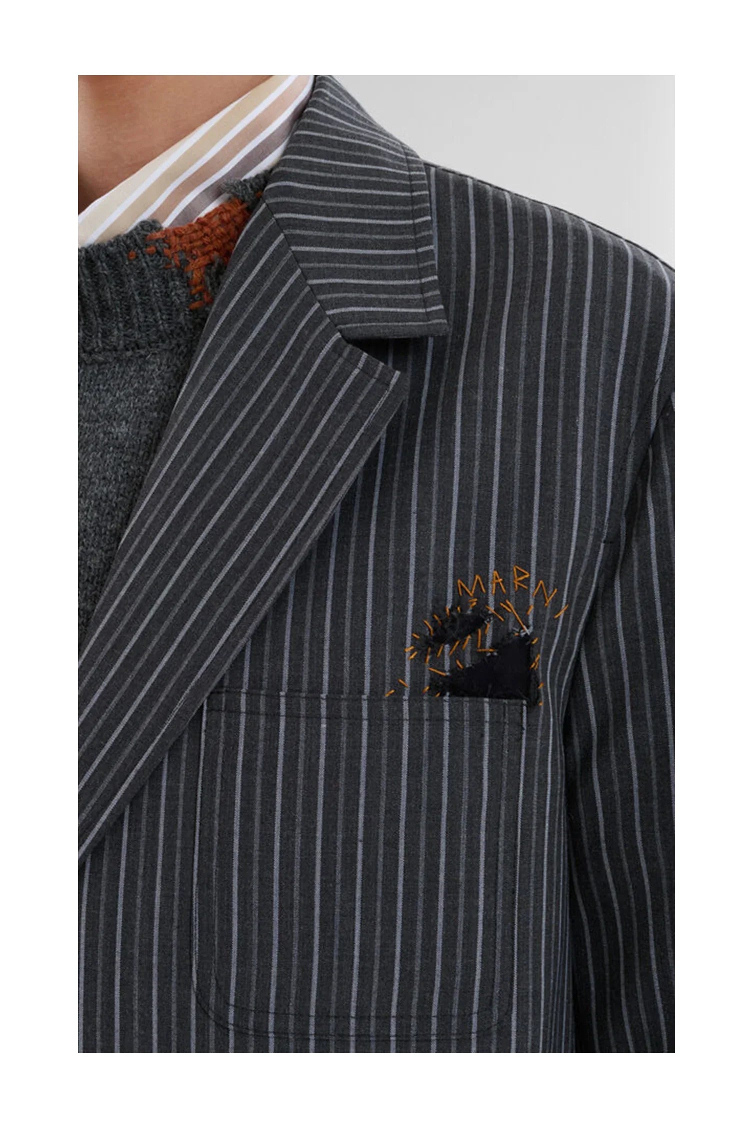 Close-up of a dark gray pinstripe blazer with embroidered pocket detail and layered knit sweater and striped shirt