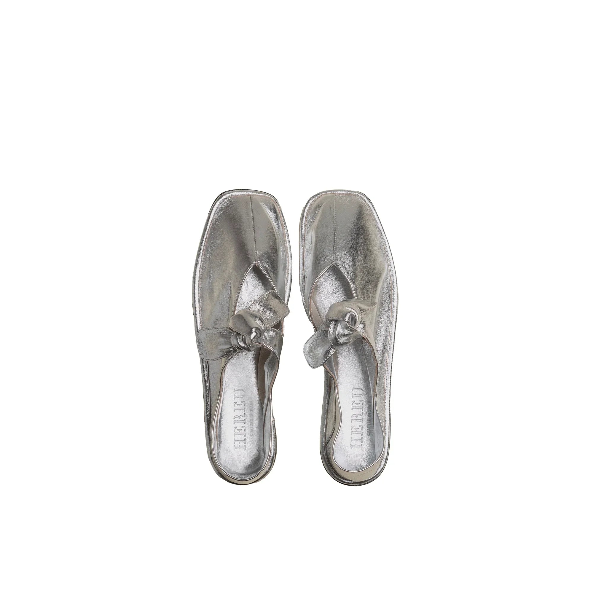 Pair of metallic silver slip-on shoes with square toes and decorative knots on white background