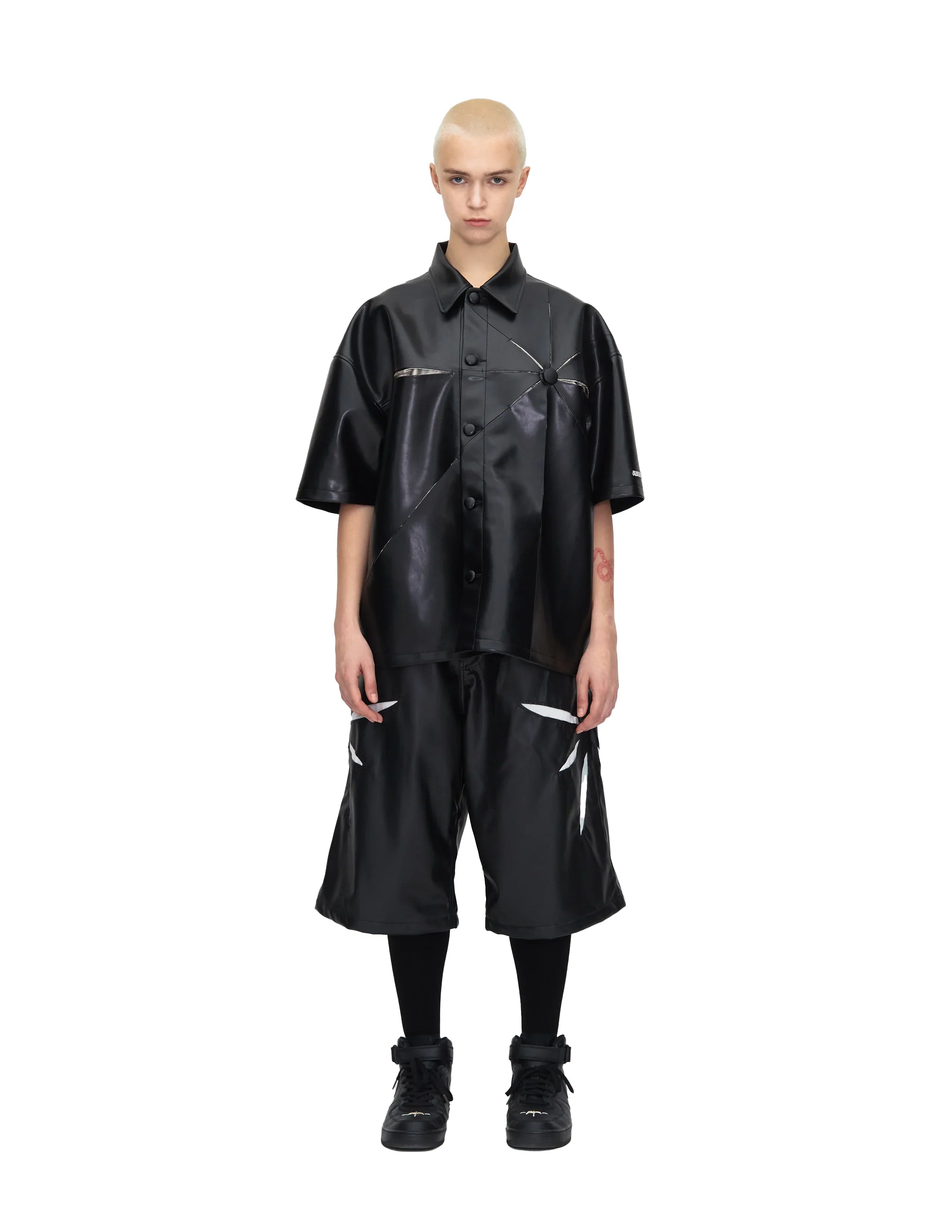 Model wearing black faux leather short-sleeve shirt and matching black long shorts with black sneakers and socks