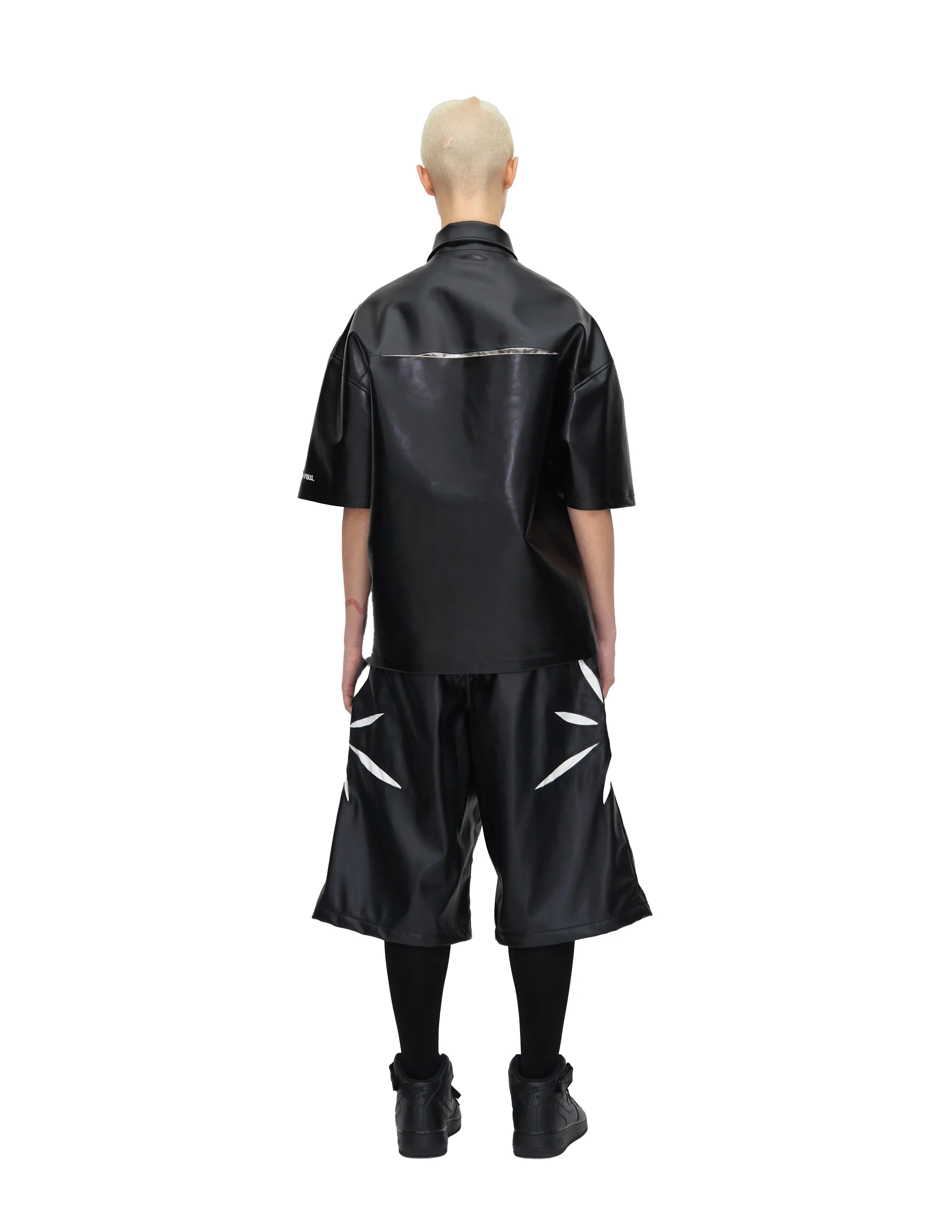 Model wearing black short sleeve leather shirt and matching wide shorts with white leaf-like design, black socks and shoes