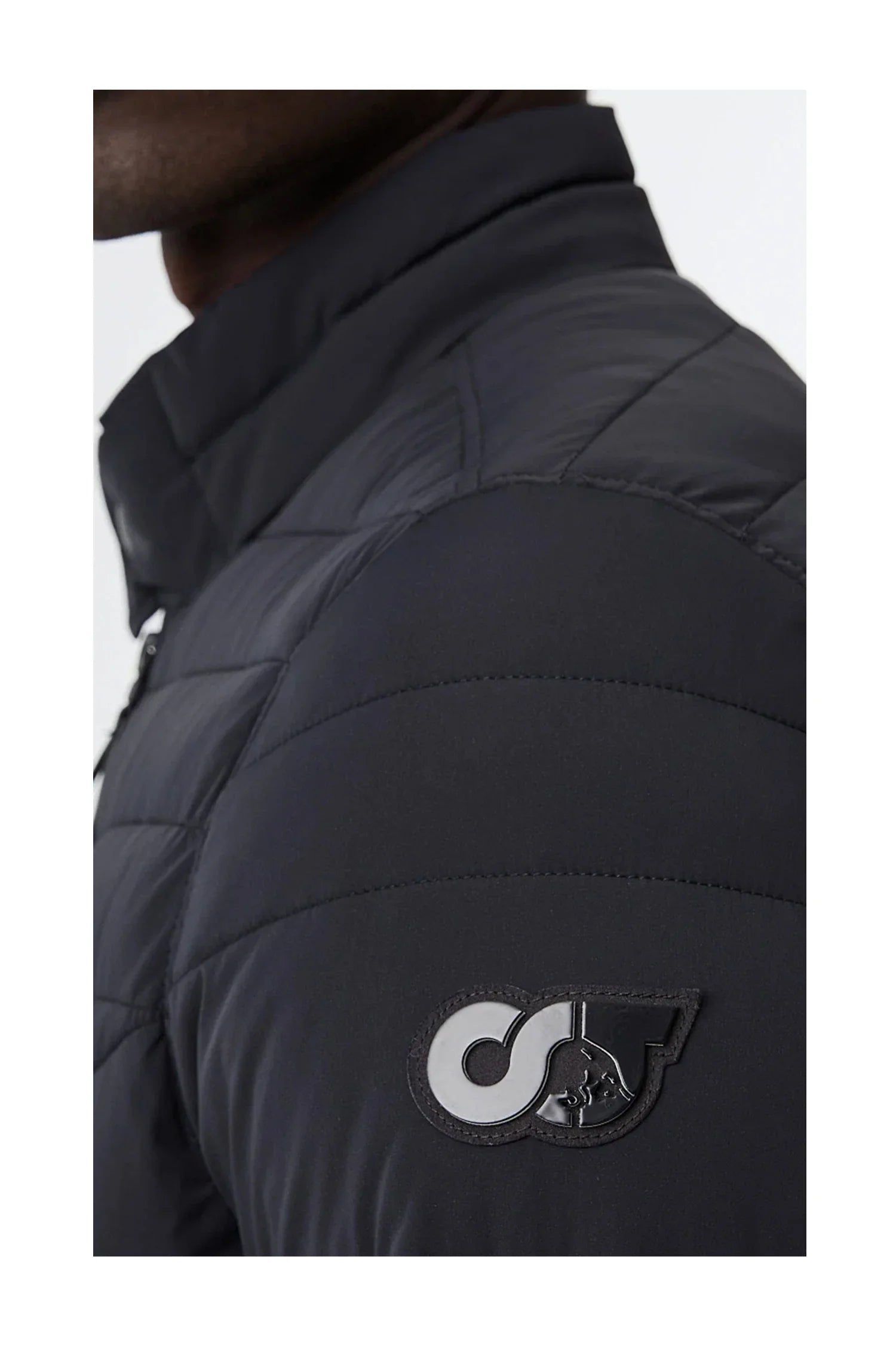 Close-up of a black quilted jacket sleeve with a stitched logo patch featuring interlocking letters and a dog silhouette
