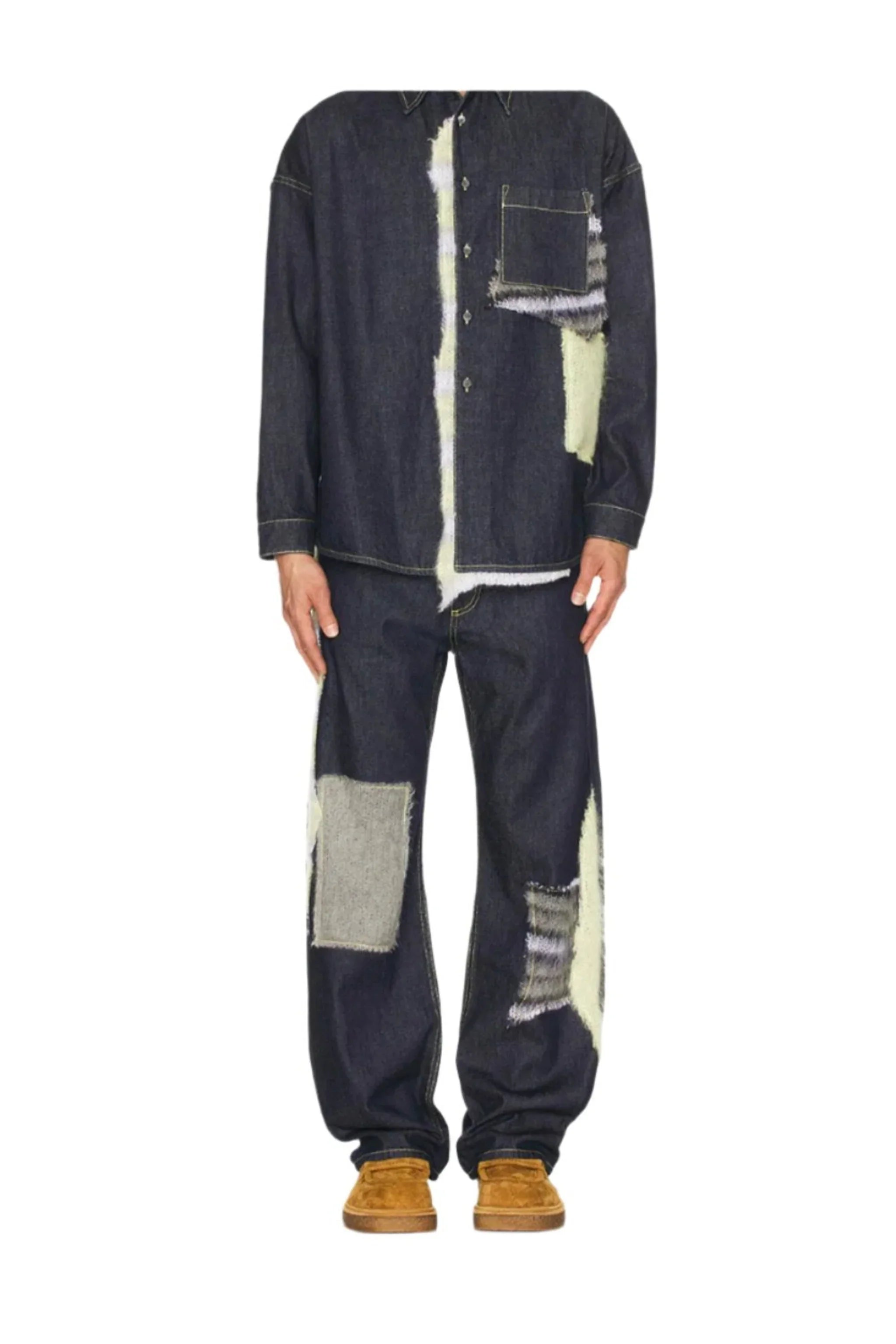 Man wearing dark denim patched shirt and pants with brown suede shoes on white background