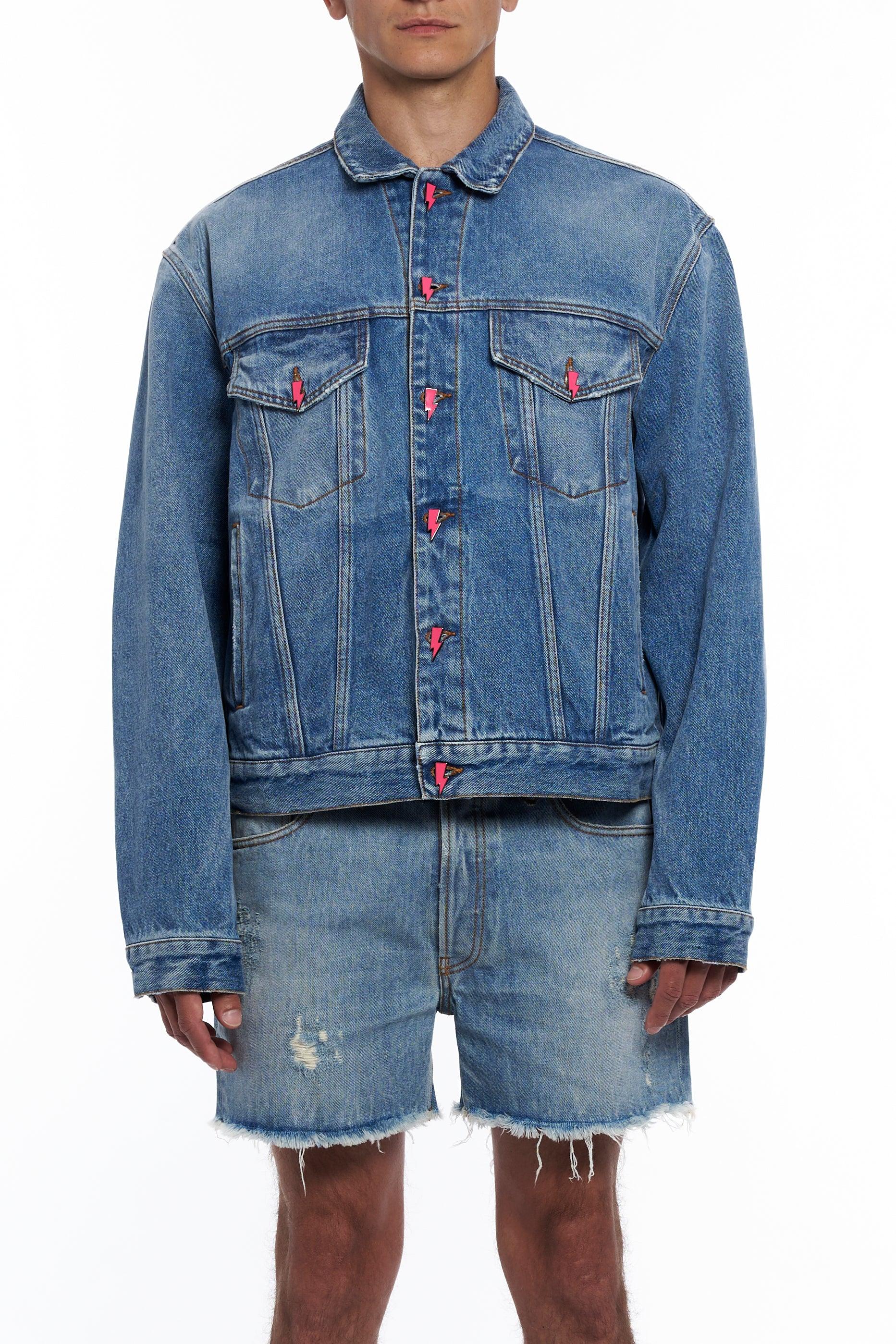 Man wearing light blue denim jacket with pink lightning bolt buttons and distressed denim shorts