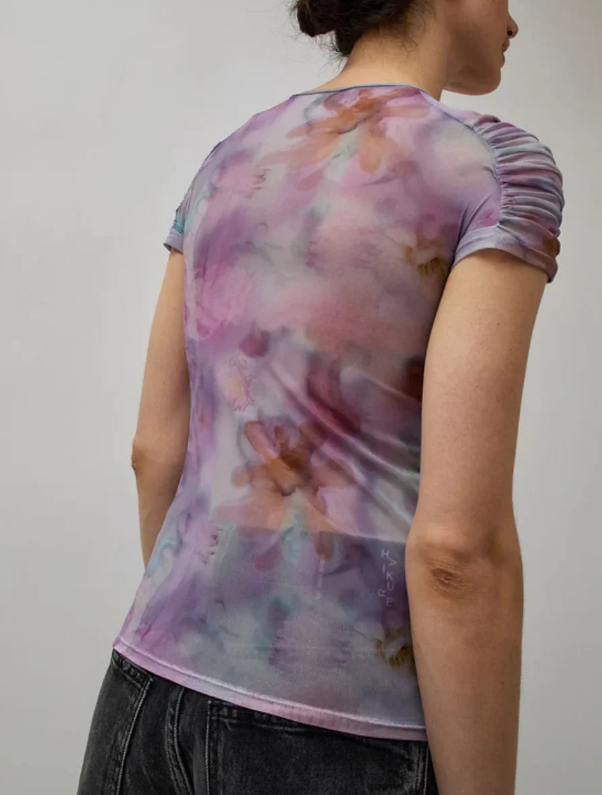 Woman wearing sheer purple floral print ruched sleeve top with black washed jeans back view