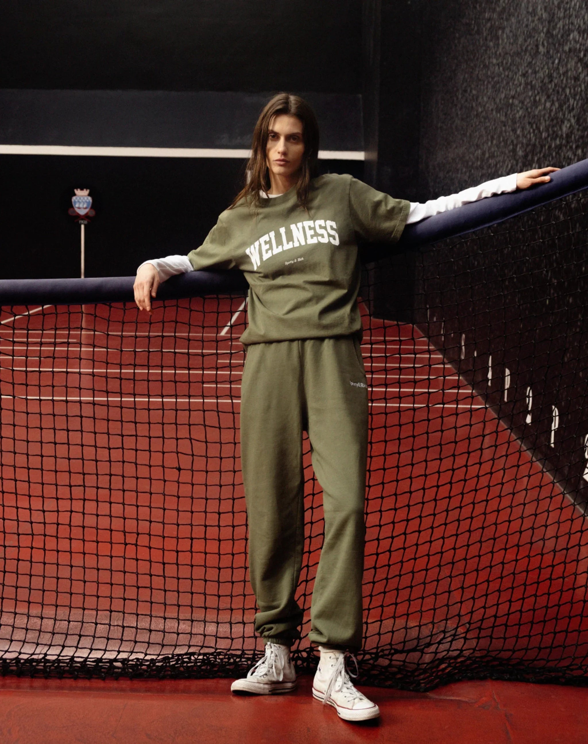 Woman in olive green wellness sweatpants and t-shirt leaning on tennis net on red indoor court