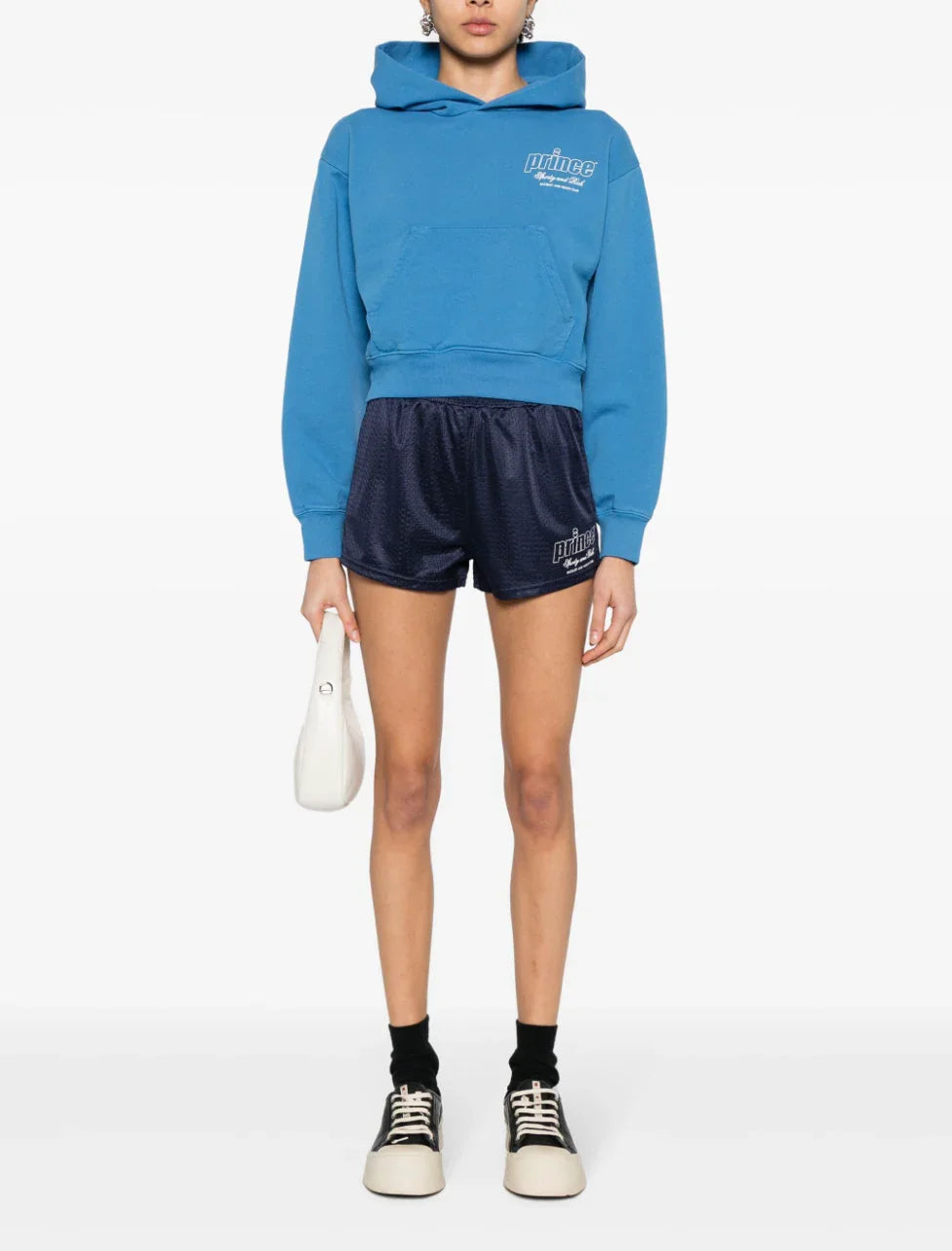 Model wearing blue cropped Prince hoodie, navy athletic shorts, black socks, sneakers, and holding white handbag