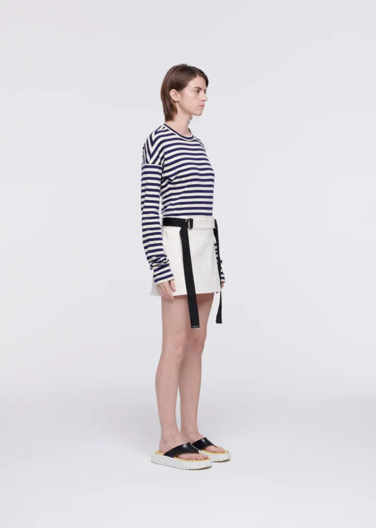 Woman in navy striped long-sleeve shirt, white mini skirt with black straps, and black slide sandals on white background