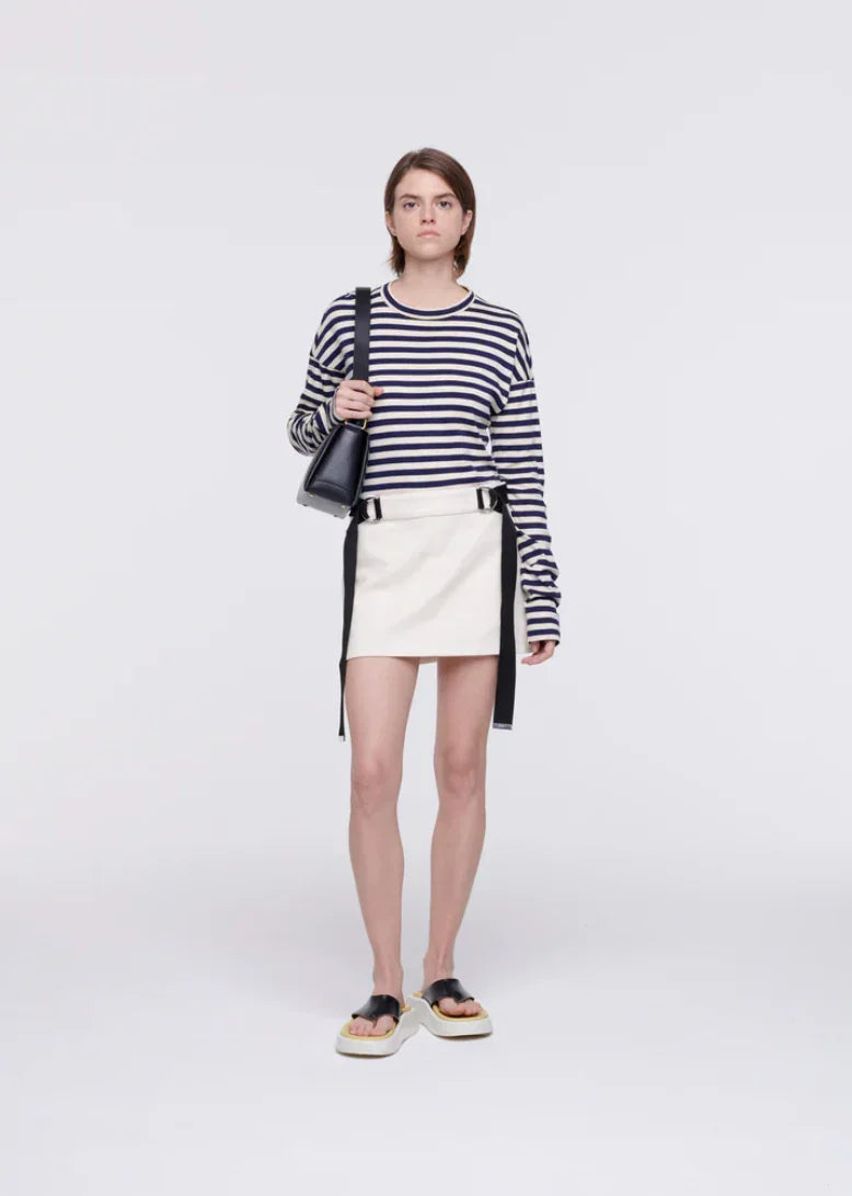 Young woman wearing navy striped long-sleeve top, white mini skirt with black straps, black sandals, and carrying black shoulder bag