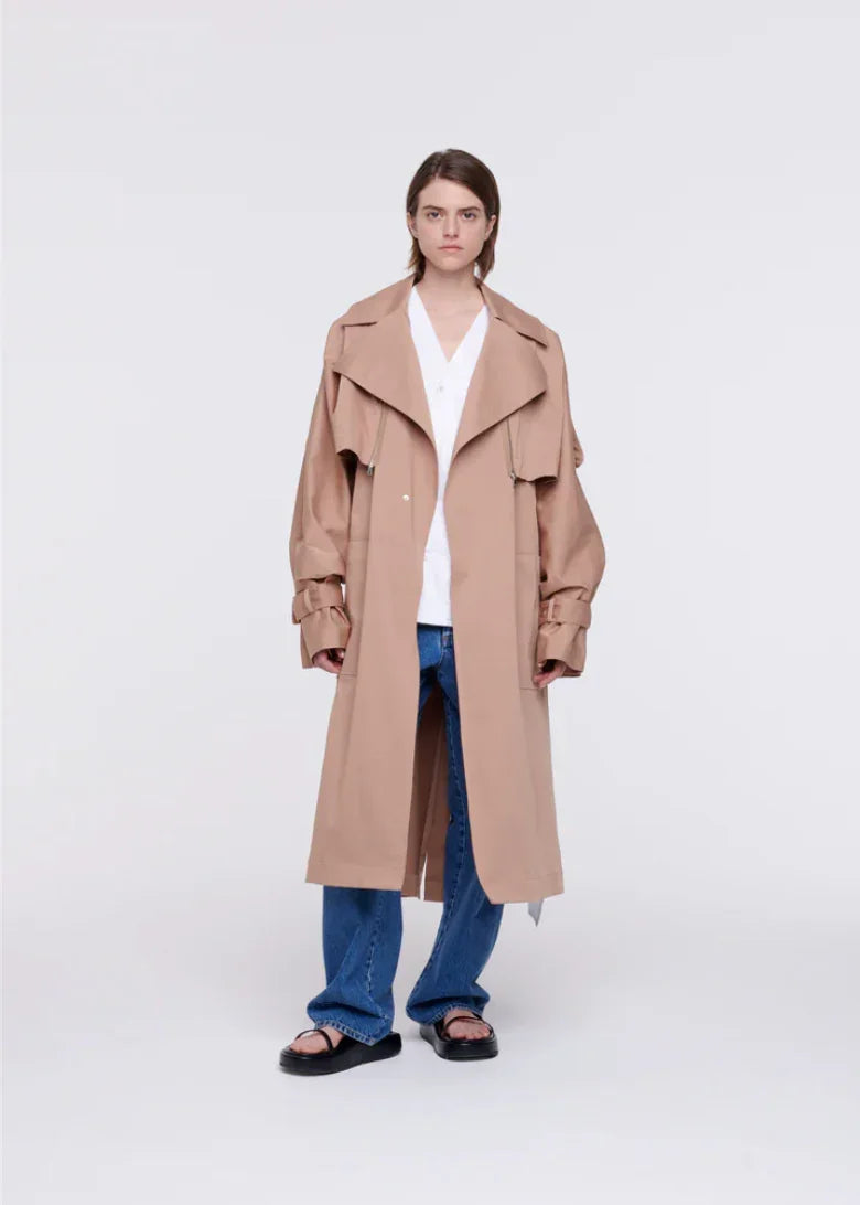 Model wearing beige oversized trench coat with blue jeans and black sandals on white background