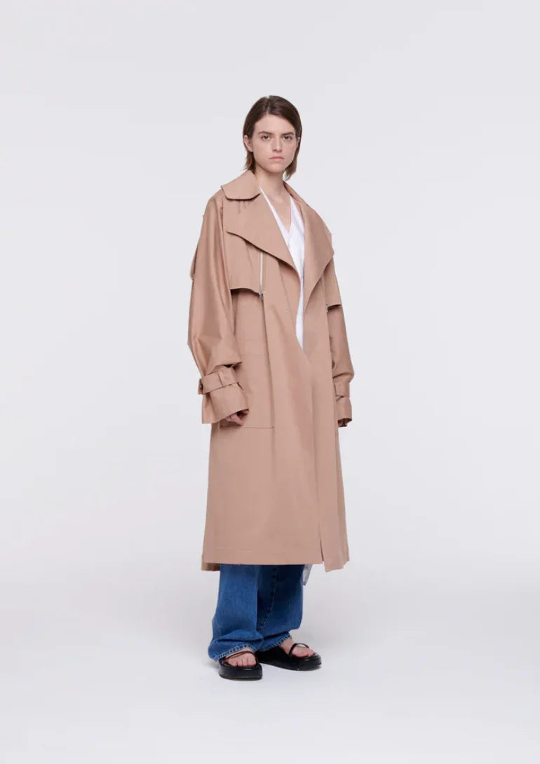 Model wearing a beige oversized trench coat with wide lapels, blue jeans, and black sandals on white background