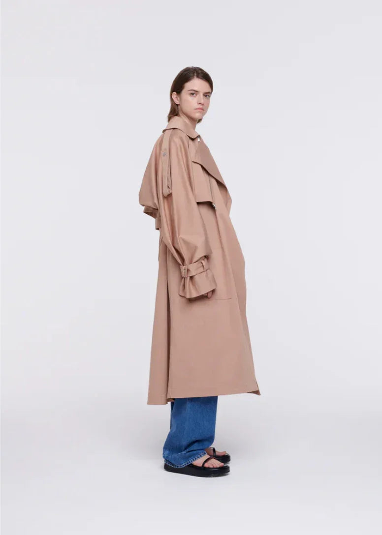 Woman wearing long beige trench coat with blue jeans and black sandals on white background