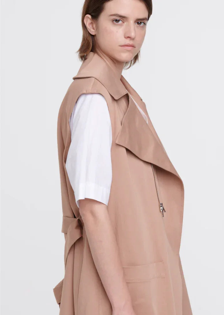 Young woman wearing beige sleeveless trench coat over white short-sleeve shirt against gray background