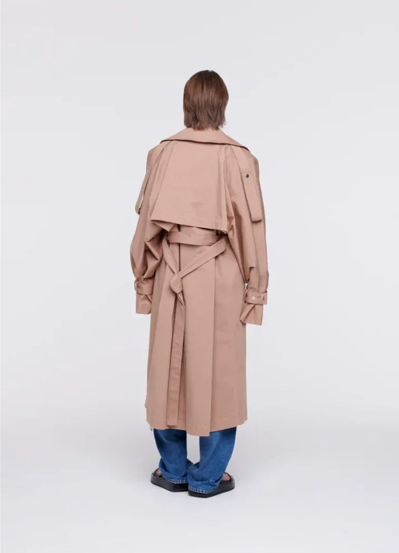 Back view of person wearing beige trench coat with belt, blue jeans, and black sandals on white background