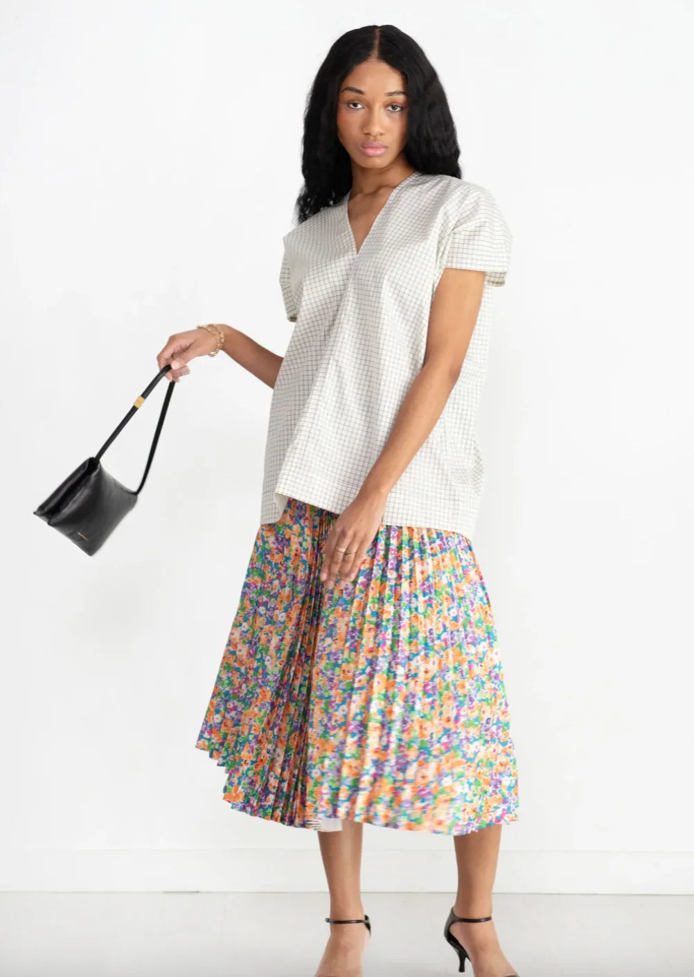 Woman wearing grid pattern V-neck top and colorful floral pleated midi skirt holding black handbag