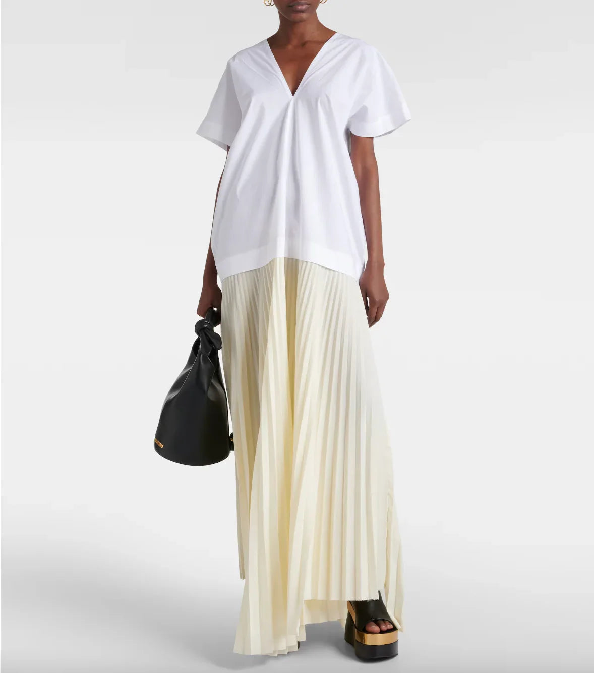 Model wearing white V-neck loose top and cream pleated maxi skirt with black platform sandals and black handbag