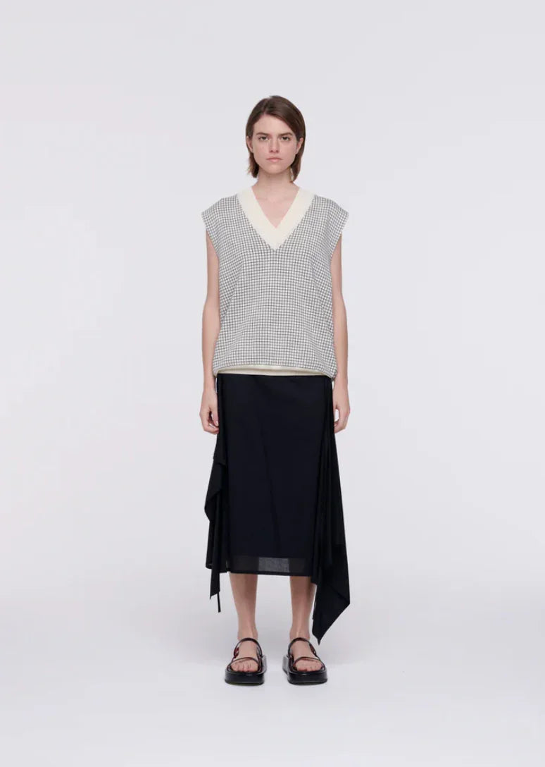Woman wearing sleeveless checkered sweater vest, black asymmetrical midi skirt, and black sandals on white background